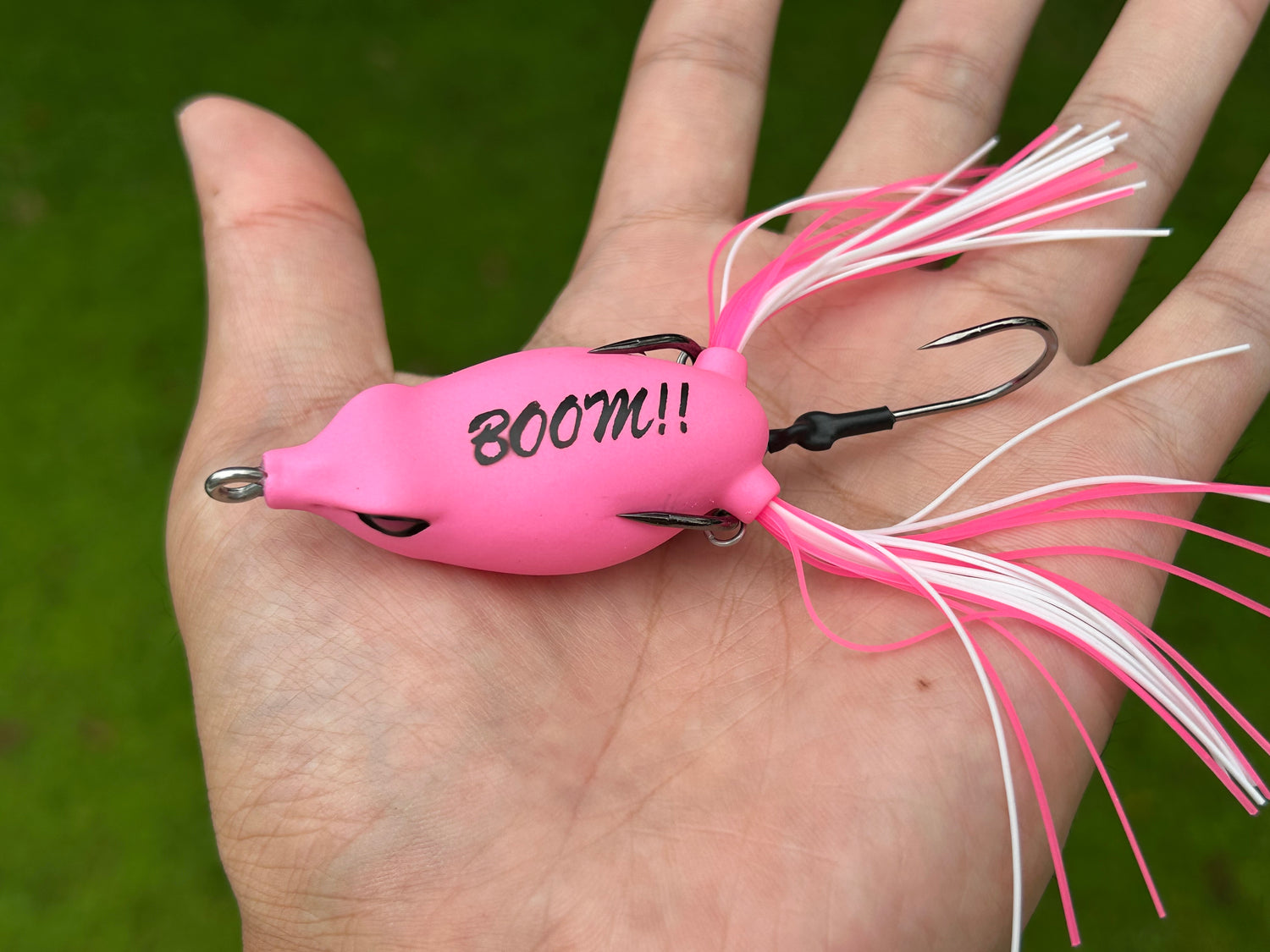 Pink fishing lure with 'BOOM!!' text held in a hand against a green background